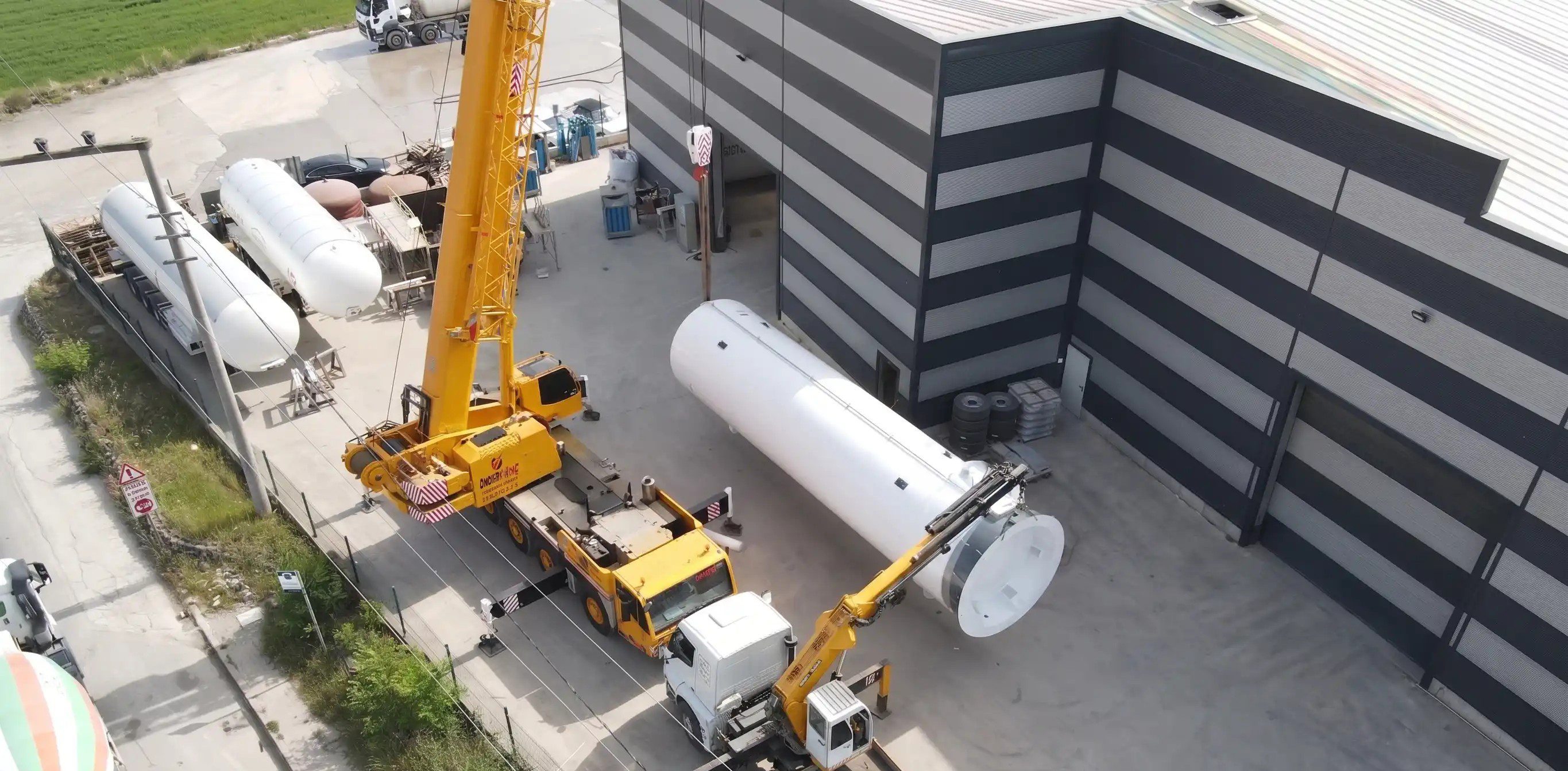 Fabrika sahasında ağır tonajlı sarı vinç yardımıyla sevkiyata hazırlanan, yüksek kapasiteli beyaz LNG depolama tankı drone görünümü.